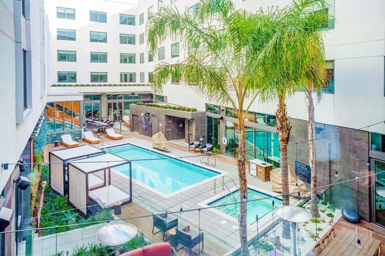 Resort courtyard with swimming pool, lounge chairs, palm trees, and modern white building