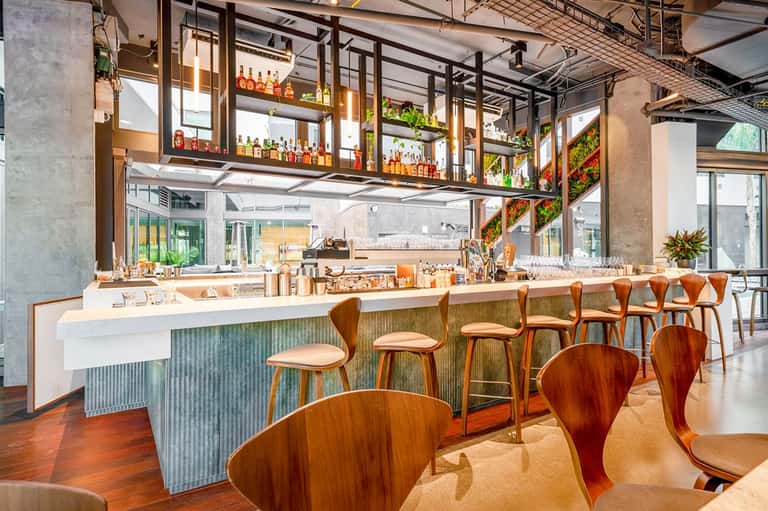 Modern bar with wooden seating, suspended bottle displays, and industrial ceiling design