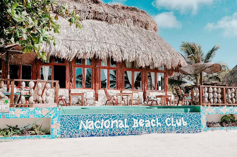 Colorful beachfront beach club with red wooden structure, thatched roof, and turquoise mosaic tile signage