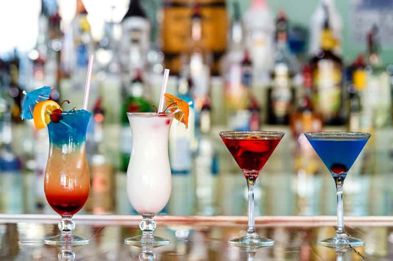 Four colorful cocktails at bar with blurred bottle collection, including blue, pink, red, and orange drinks