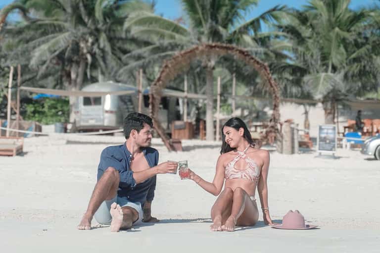 Couple toasting drinks on a sandy beach with palm trees and beach structures in the background