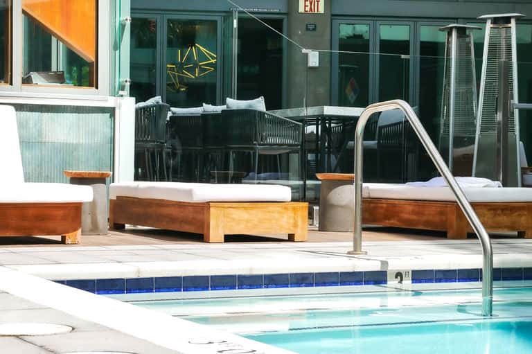 Modern hotel pool with wooden lounge chairs, metal railings, and glass building with green doors