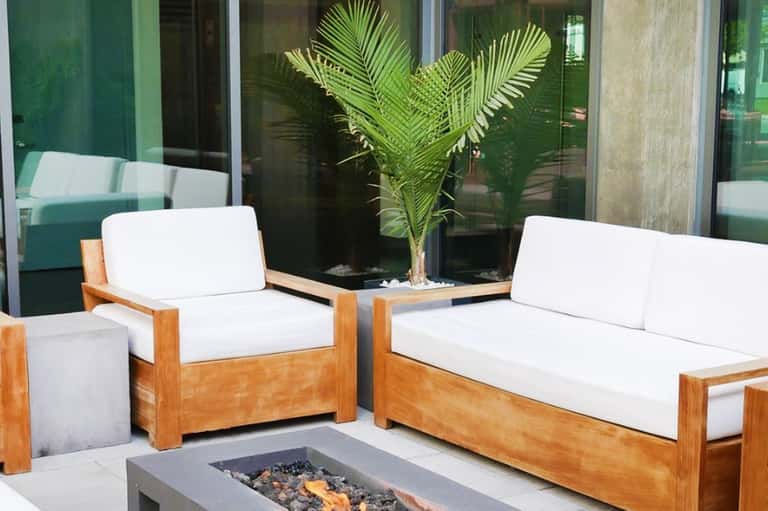 Modern outdoor lounge seating with white cushions, wooden frames, potted palm plant, and fire pit