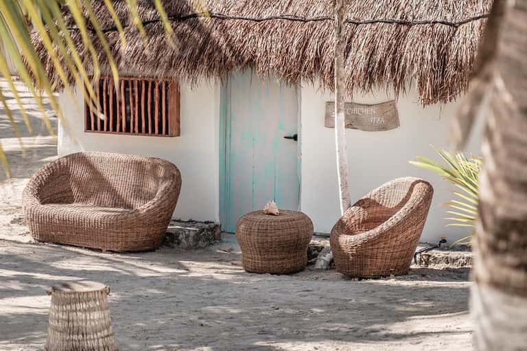 Woven rattan chairs on sand outside a white bungalow with thatched roof