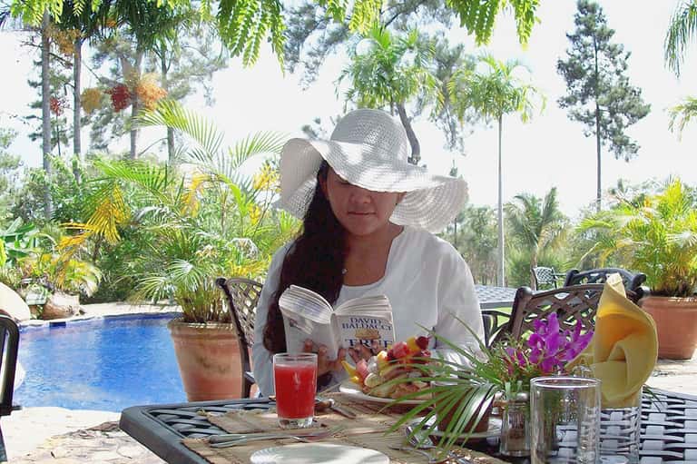 Woman reading poolside in tropical setting with fresh beverages, flowers, and lush garden landscape