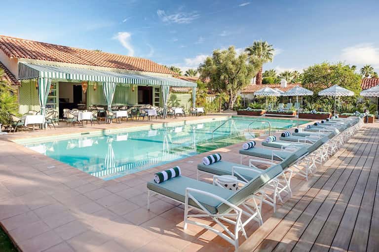 Resort pool with white lounge chairs, striped cabana, tropical landscaping, and palm trees under blue sky