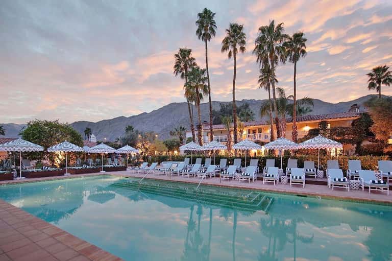 Desert resort pool at dusk with palm trees, umbrellas, and illuminated buildings
