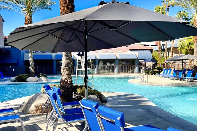 Resort pool area with large black umbrella, blue lounge chairs, palm trees, and modern buildings