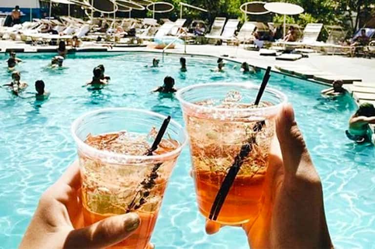 Two iced beverages held up at resort pool with swimmers and lounge chairs visible in background