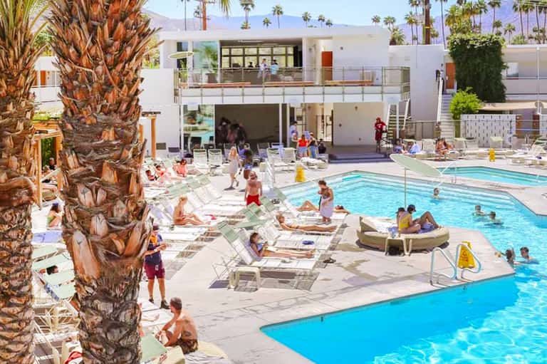 Resort pool deck with guests lounging on chairs, palm trees, modern white building, and mountains in distance