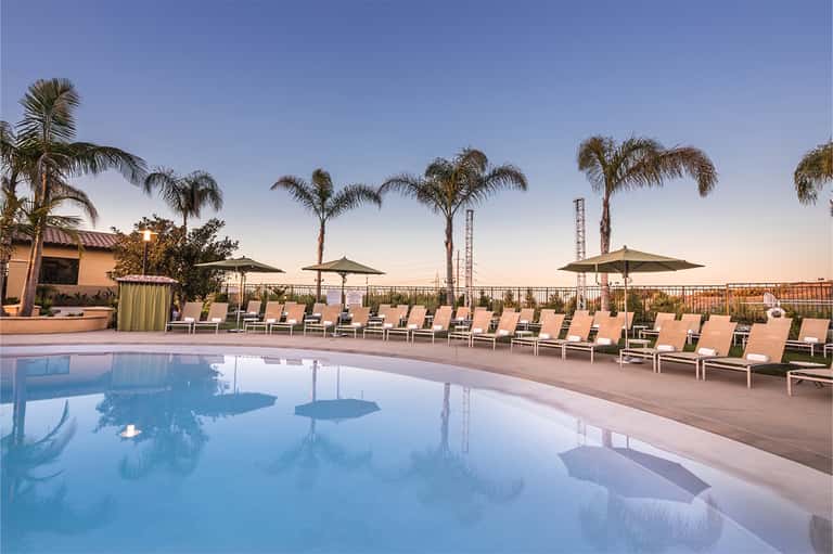 Resort pool at dusk with palm trees, lounge chairs, umbrellas, and amusement park rides visible