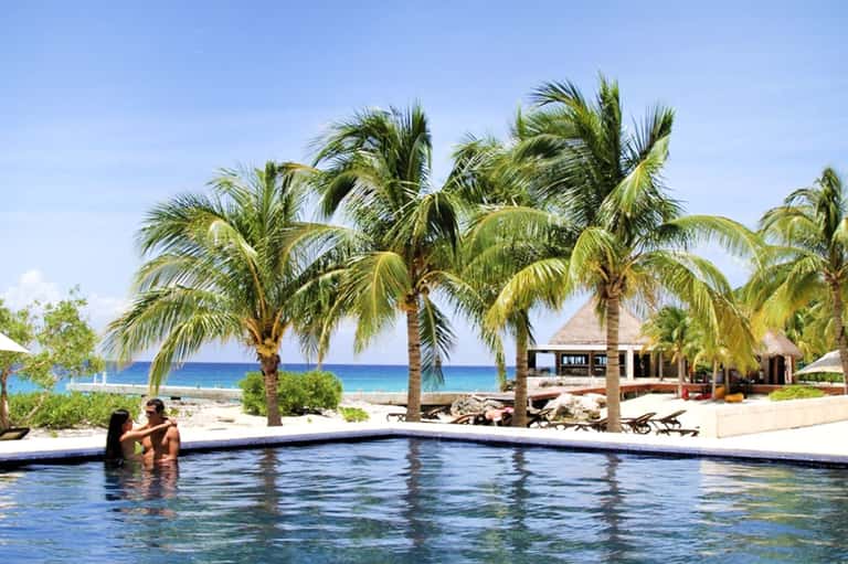 Couple wading in a tropical resort infinity pool with palm trees and beach bungalows visible