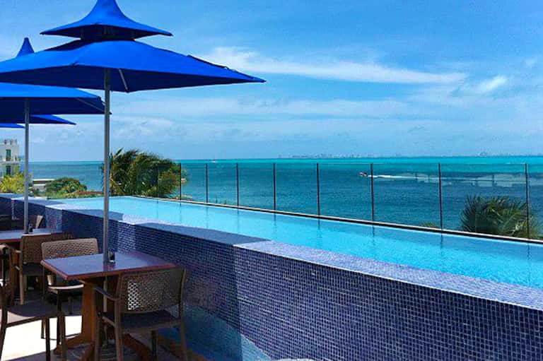 Modern resort infinity pool overlooking turquoise ocean with blue umbrellas, dining furniture, and coastal landscape