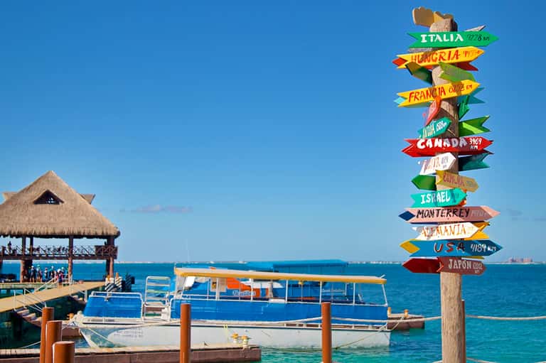 Directional signpost with international city names at a tropical dock with boats and thatched bar