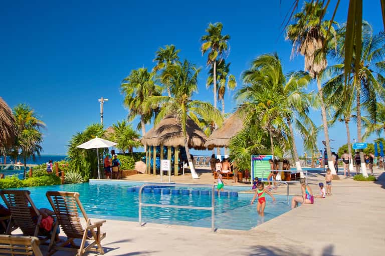 Family-friendly resort pool with palm trees, thatched roof structures, and children swimming