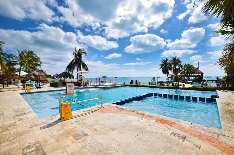 Resort swimming pool with palm trees, tiki huts, beach access, and clear Caribbean waters on sunny day