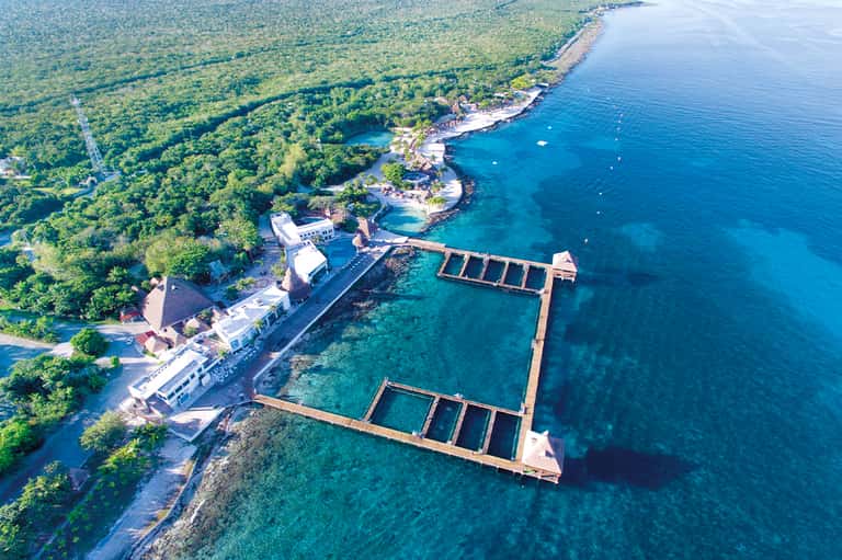 Aerial view of oceanfront resort with natural pools, wooden docks, lush tropical forest, and turquoise bay waters