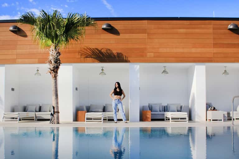 Woman standing by pool in modern resort with white cabanas and wooden pergola