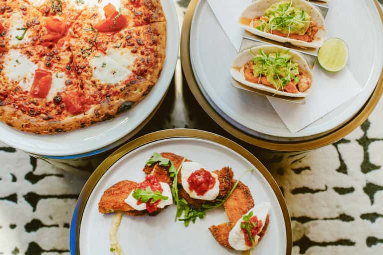 Overhead view of gourmet dishes: pizza, tacos, and fried appetizers on white and tan rimmed plates