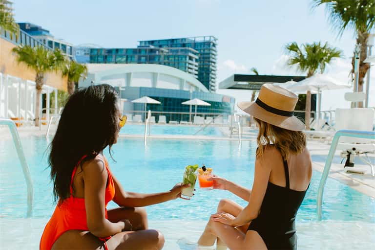 Two women toasting colorful cocktails by resort pool with palm trees and modern buildings