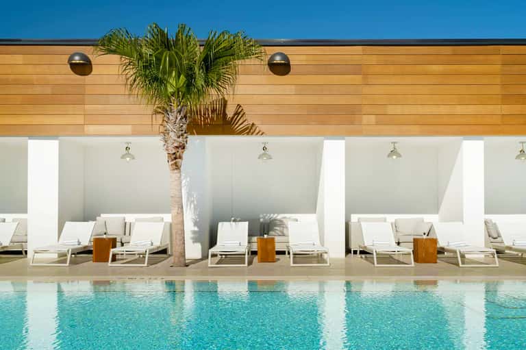 Resort poolside with white loungers, palm tree, wooden ceiling panels, and clear blue pool