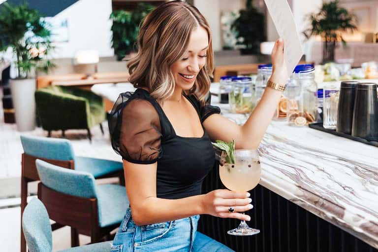 Woman in black top at elegant resort bar holding a cocktail and smiling at the camera