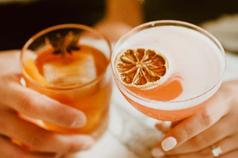 Two cocktails with dried citrus garnish held in hands at sunset