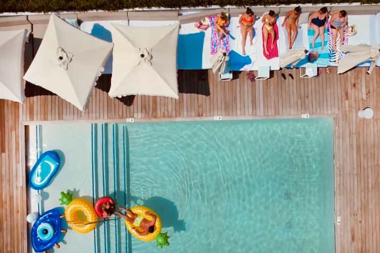 Overhead view of pool with people sunbathing, float toys, umbrellas, and wooden deck beside water