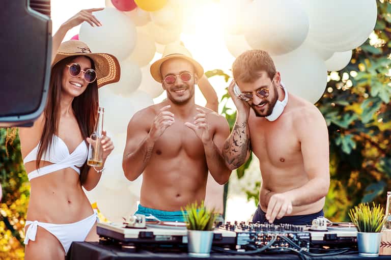 Three people at an outdoor DJ booth with white balloons and tropical foliage
