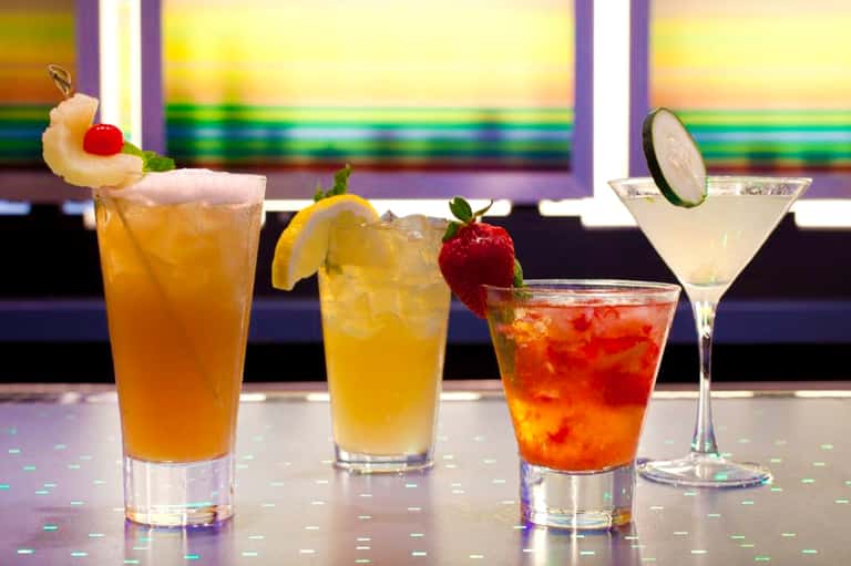 Colorful tropical cocktails with fruit garnishes displayed on counter with rainbow-striped backdrop