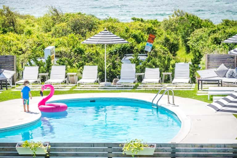Resort swimming pool with pink flamingo float, white lounge chairs, striped umbrellas, and ocean view beyond vegetation