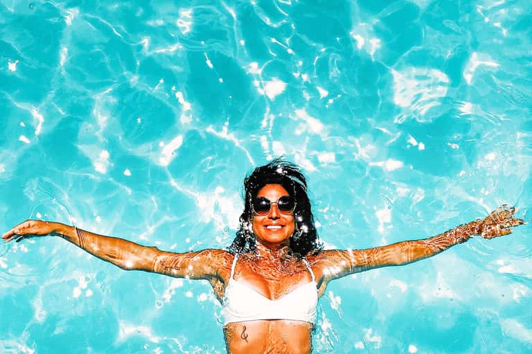 Woman swimming in bright turquoise pool with arms spread wide, wearing sunglasses and white top