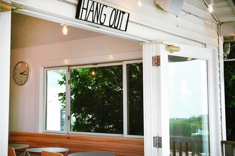 Modern outdoor café or restaurant space with 'HANG OUT' sign, clock, and glass doors overlooking greenery