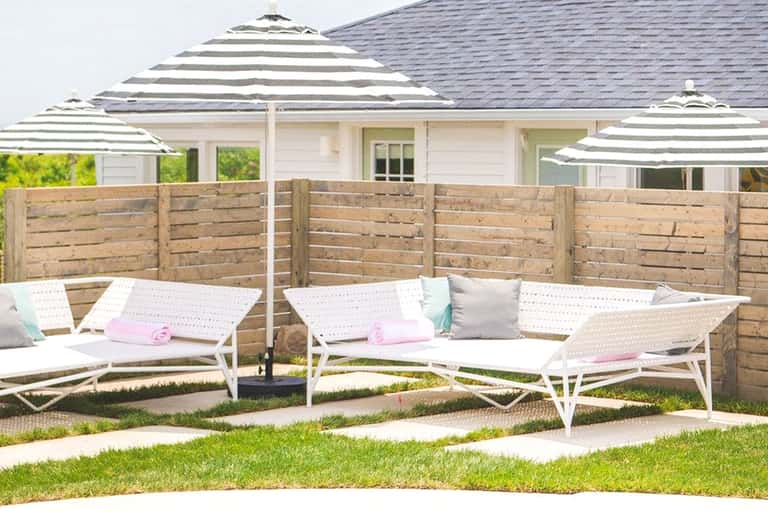 Residential backyard patio with striped umbrellas, white loungers, and wooden fence