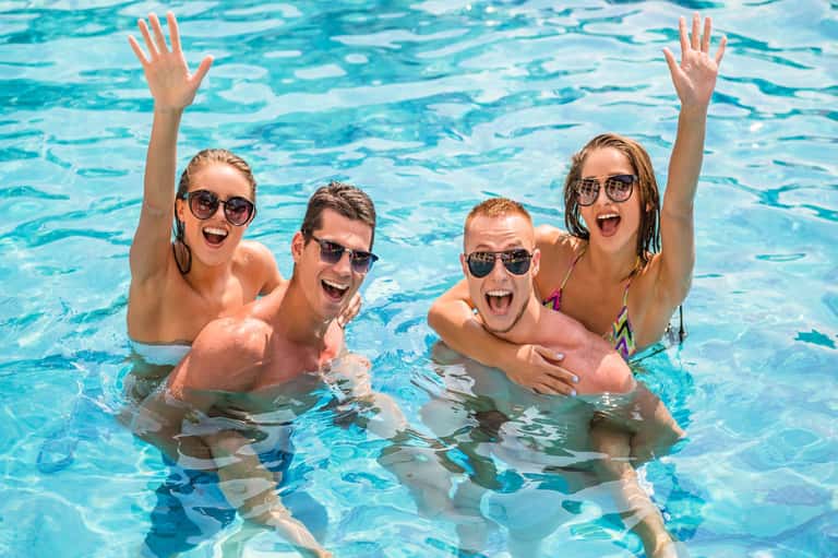 Four young adults enjoying themselves in a bright blue pool, smiling and raising hands