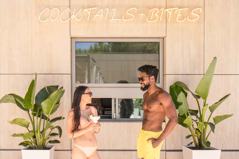 Couple at poolside cocktails bar with neon sign, tropical plants, and modern architecture