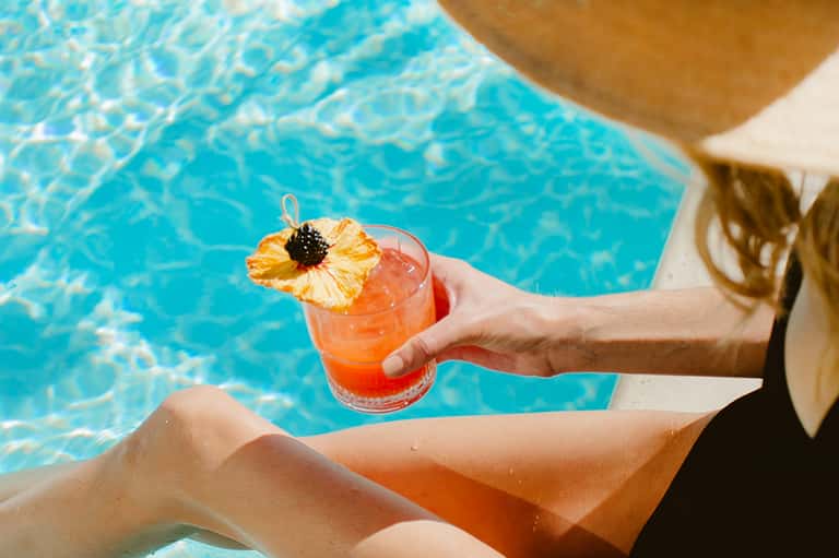 Person holding a colorful tropical cocktail with pineapple garnish overlooking a bright blue swimming pool