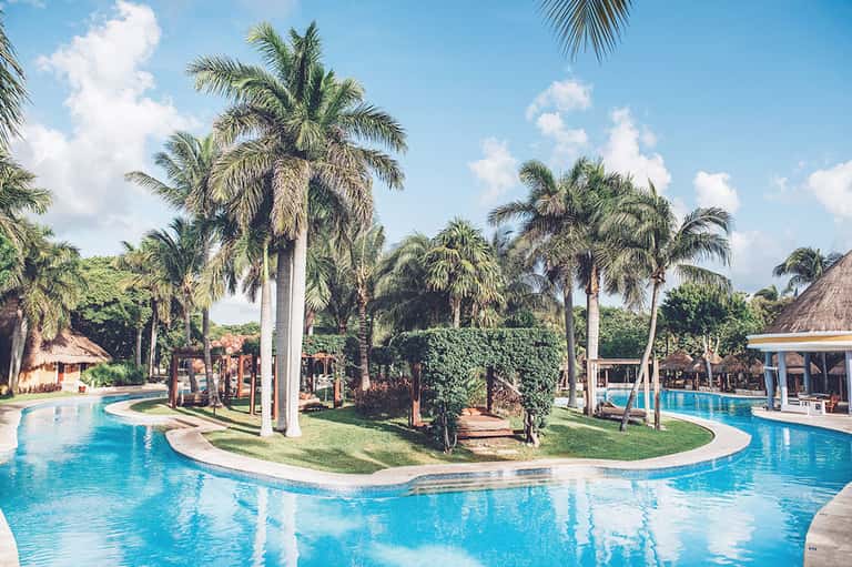 Resort swimming pool with curved design, palm trees, tiki huts, and manicured lawn area