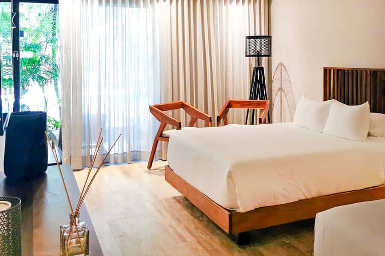 Bright hotel bedroom with white bedding, modern wood furniture, and balcony overlooking tropical landscape