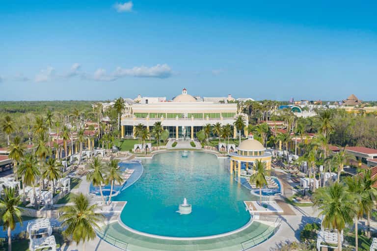 Aerial view of resort with curved turquoise pool, palm-lined grounds, and main building