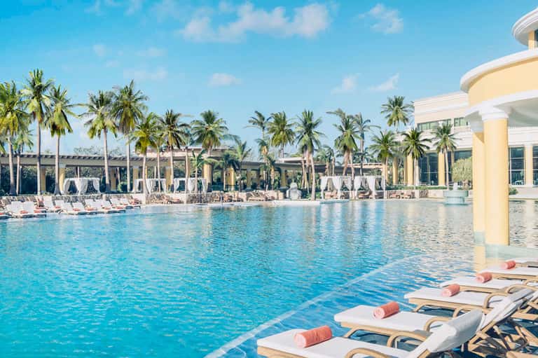 Resort lagoon pool surrounded by palm trees, lounge chairs, and cream-colored building architecture