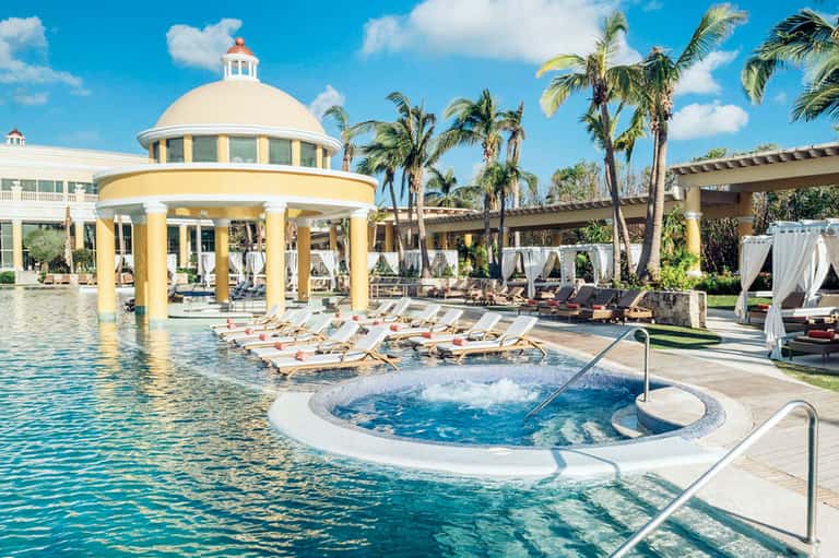 Resort pool with yellow cabana, lounge chairs, spa tub, and palm trees with clear blue sky