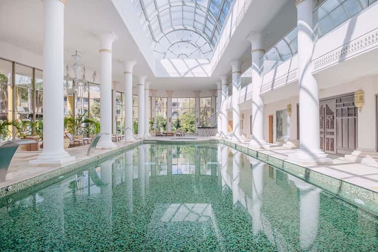 Indoor lap pool with glass ceiling, white columns, and surrounding lounge seating areas