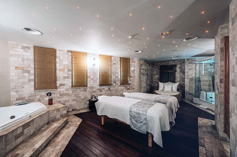 Luxury spa suite with two beds, marble walls, soaking tub, heated mirrors, and dark wood flooring