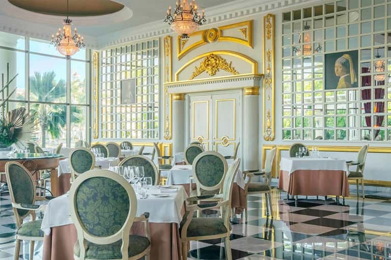 Elegant fine dining restaurant with white tablecloths, gold ornate décor, and palm view windows
