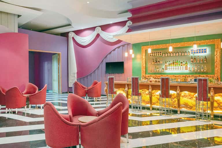 Vibrant hotel bar lounge with pink curved chairs, ornate gold mirror, colorful ceiling details, and bar seating