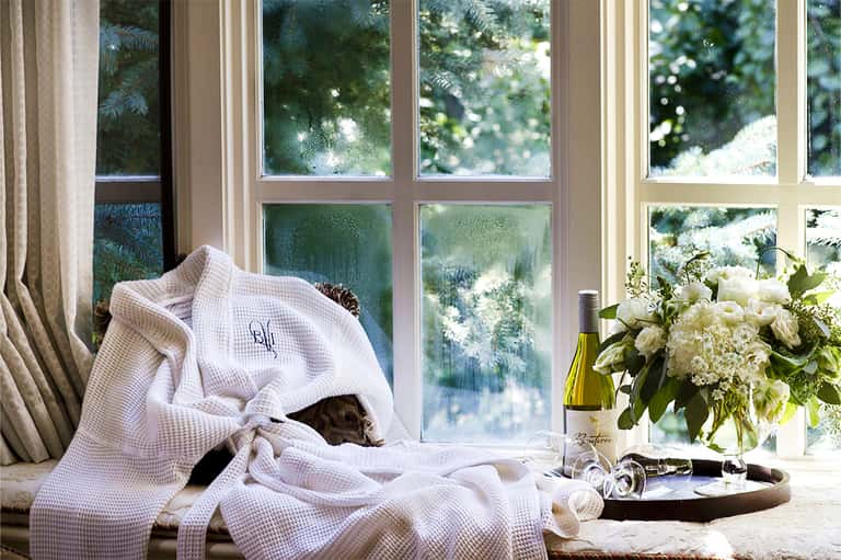 Luxury bedroom window seat with white robe, flowers, wine bottle, and garden views