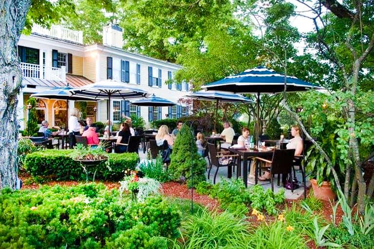 Outdoor dining area under blue umbrellas near colonial-style white house with gardens