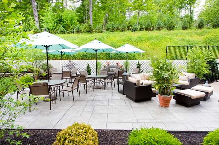 Upscale backyard patio with white umbrellas, wicker lounge furniture, and manicured landscape with forest views