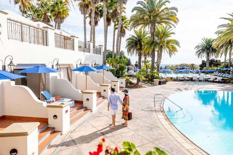 Beachfront pool area with cabanas, blue umbrellas, and palm trees lining the shore